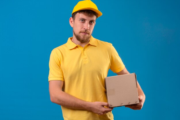 entregador de camisa pólo amarela e boné segurando um pacote de caixa parecendo incerto de pé no azul