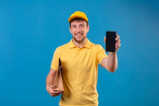 entregador de camisa pólo amarela e boné segurando um pacote de papel e mostrando o smartphone sorrindo amigável com uma cara feliz em pé no azul