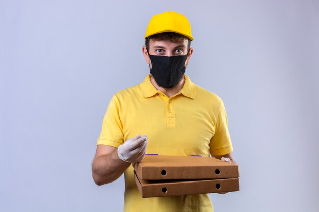 Entregador de camisa pólo amarela e boné usando máscara protetora preta com sorriso no rosto fazendo gesto de dinheiro com a mão em pé no branco