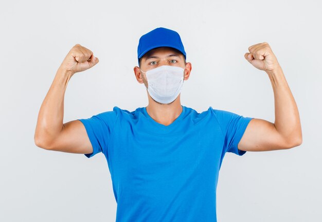 Entregador de camiseta azul, boné e máscara mostrando os músculos e parecendo forte