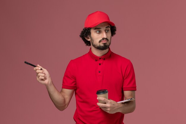 Entregador de correio masculino de camisa vermelha e capa, vista frontal, segurando a xícara de café marrom e o bloco de notas com caneta na parede rosa claro.