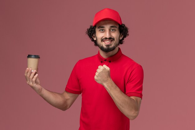 Entregador de correio masculino de camisa vermelha e capa, vista frontal, segurando a xícara de café marrom na parede rosa serviço uniforme empregado de entrega trabalho masculino