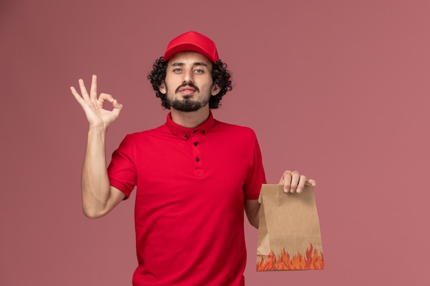 Entregador de correio masculino de camisa vermelha e capa, vista frontal, segurando um pacote de comida na parede rosa.