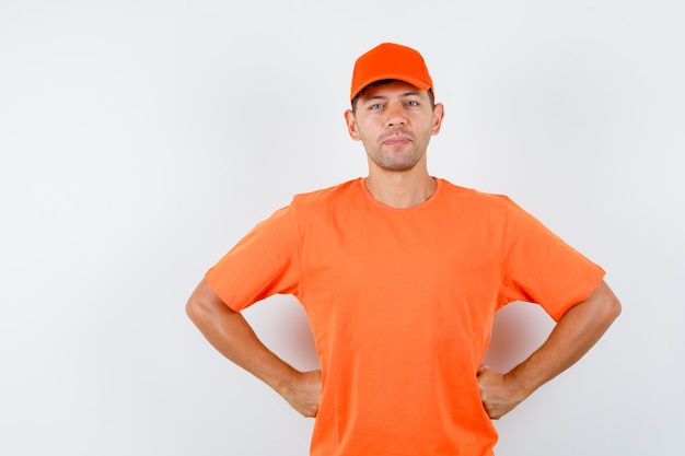 Entregador de mãos dadas na cintura e sorrindo em vista frontal de t-shirt e boné laranja.