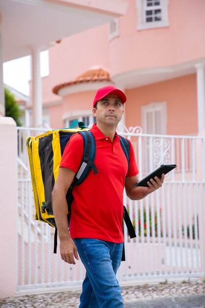 Entregador de meia-idade olhando para longe, carregando bolsa térmica e segurando o tablet. Correio caucasiano de camisa vermelha e boné, caminhando pela rua e entregando o pedido. Serviço de entrega e pós-conceito