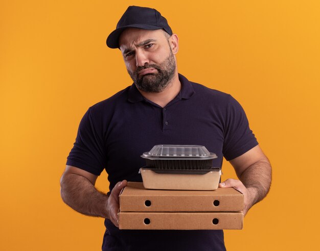 Entregador de meia-idade, triste, de uniforme e boné segurando um recipiente de comida em caixas de pizza isoladas na parede amarela