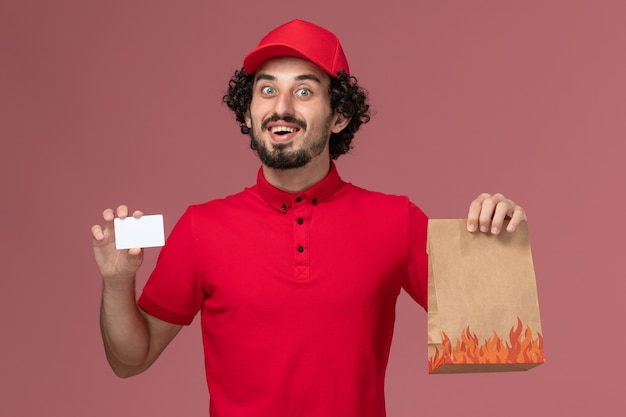 Entregador de mensageiro masculino de camisa vermelha e capa de frente segurando um pacote de comida e cartão na parede rosa.