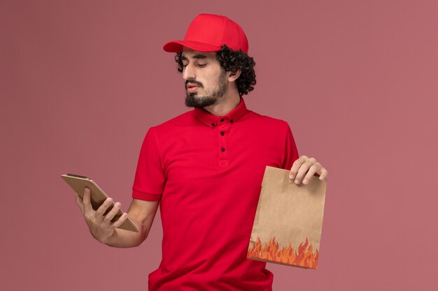 Entregador de mensageiro masculino de camisa vermelha e capa de frente segurando um pacote de comida e o bloco de notas na parede rosa.