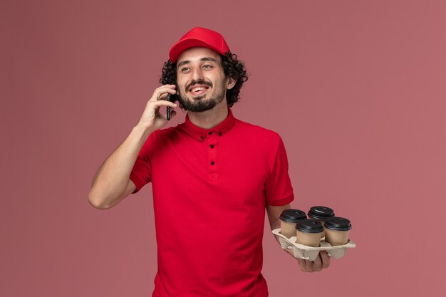 Entregador de mensageiro masculino de camisa vermelha e capa segurando xícaras de café de frente, falando ao telefone na parede rosa claro trabalho de funcionário de entrega de serviço masculino