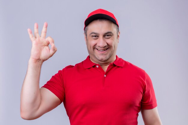 Entregador de uniforme vermelho e boné parecendo positivo e feliz, sorrindo alegremente fazendo sinal de ok em pé sobre um espaço em branco isolado