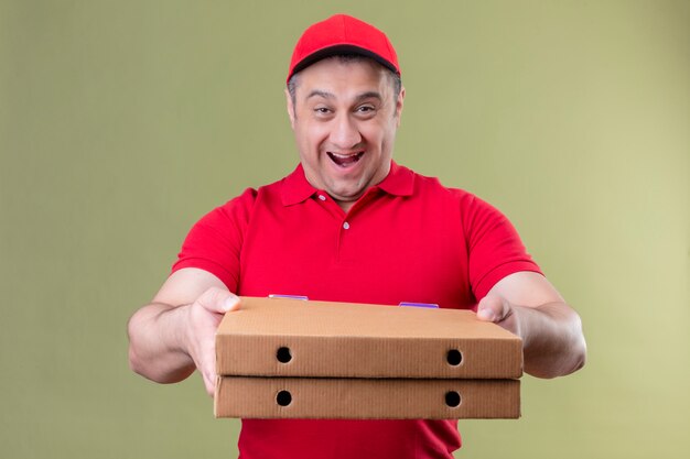 Entregador de uniforme vermelho e boné segurando caixas de pizza esticando-se para a câmera sorrindo alegremente com uma cara feliz em pé no verde isolado