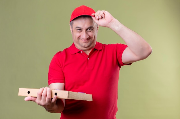 Foto grátis entregador de uniforme vermelho e boné segurando caixas de pizza positivas e feliz tocando seu boné sorrindo amigável em pé sobre o espaço verde isolado