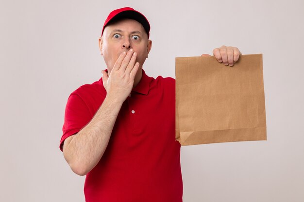 Entregador de uniforme vermelho e boné segurando um pacote de papel olhando para a câmera sendo shocekd cobrindo a boca com a mão em pé sobre um fundo branco
