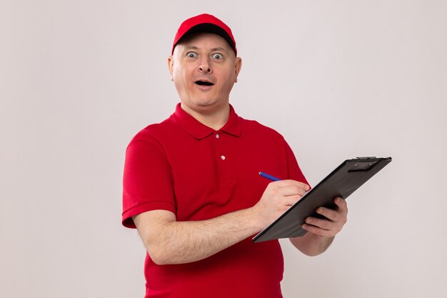 Entregador de uniforme vermelho e boné segurando uma prancheta e uma caneta, fazendo anotações olhando para a câmera feliz e animado em pé sobre um fundo branco