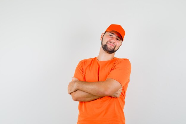 Entregador em pé com os braços cruzados em camiseta laranja, boné e parecendo alegre. vista frontal.