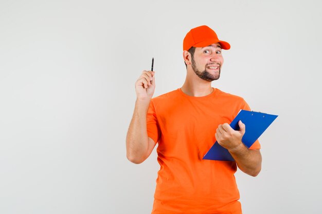 Entregador em t-shirt laranja, boné segurando a prancheta e caneta e olhando alegre, vista frontal.