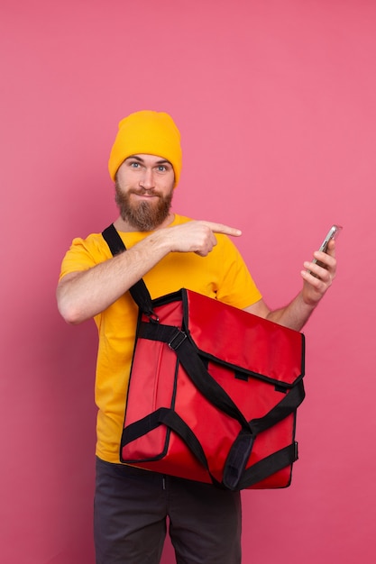 Entregador europeu alegre com bolsa casual segurando telefone apontando dedo na tela rosa