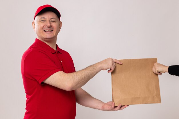 Entregador feliz e positivo em uniforme vermelho e boné dando um pacote de papel para o cliente sorrindo amigável