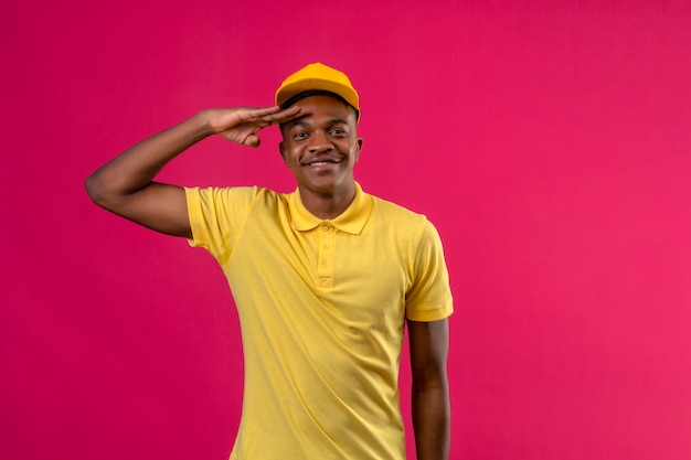 Foto grátis entregador homem afro-americano de camisa pólo amarela e boné saudando, parecendo confiante, sorrindo em pé na rosa isolada