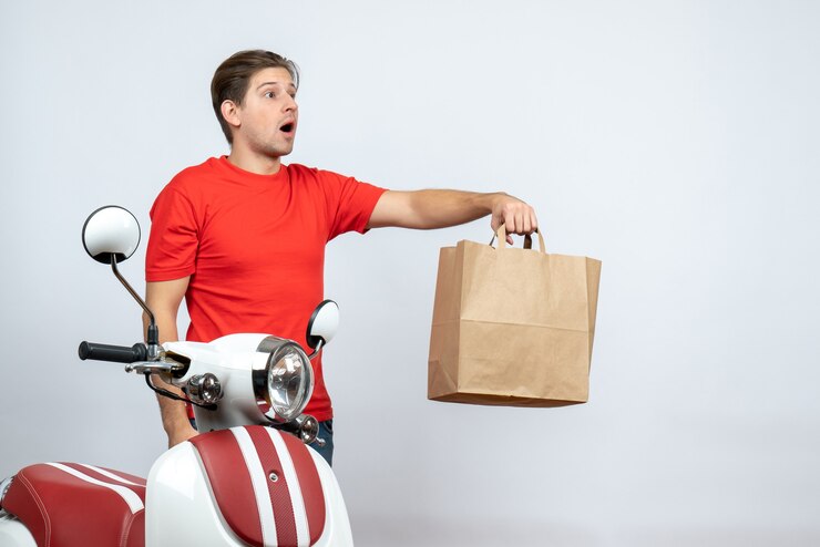 Foto grátis entregador inseguro de uniforme vermelho em pé perto da scooter dando uma caixa de papel na parede