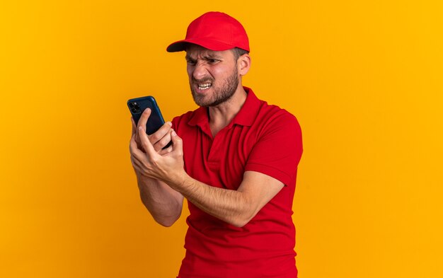 Entregador jovem agressivo, caucasiano, de uniforme vermelho e boné em pé na vista de perfil, segurando e olhando para o telefone celular isolado na parede laranja com espaço de cópia