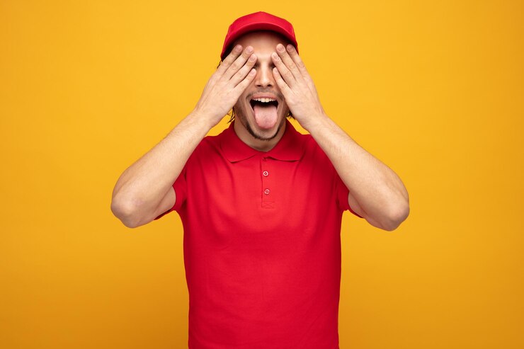 Foto grátis entregador jovem brincalhão vestindo uniforme e boné fechando os olhos com as mãos mostrando a língua isolada em fundo amarelo