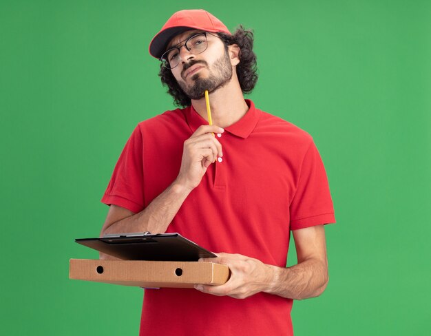 Entregador, jovem, caucasiano, confuso, de uniforme vermelho e boné de óculos, segurando a prancheta de pacote de pizza, tocando o queixo com um lápis