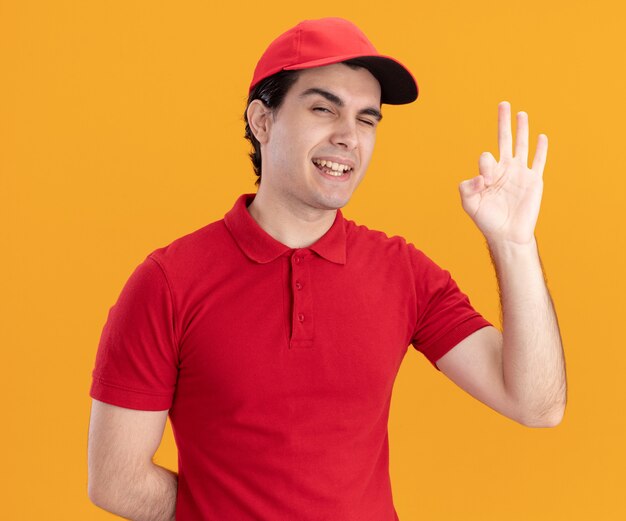 Entregador jovem confiante, caucasiano, de uniforme azul e boné, mantendo a mão atrás das costas, piscando e fazendo sinal de ok isolado na parede laranja