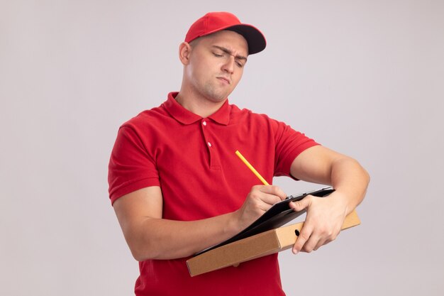 Entregador jovem confiante vestindo uniforme com tampa segurando uma caixa de pizza e escrevendo algo na prancheta, isolada na parede branca