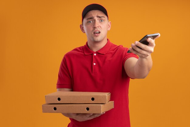 Entregador jovem confuso, vestindo uniforme com tampa segurando caixas de pizza e segurando o telefone na frente, isolado na parede laranja