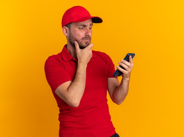 Entregador, jovem, duvidoso, caucasiano, de uniforme vermelho e boné, segurando o queixo, segurando e olhando para o celular isolado na parede laranja