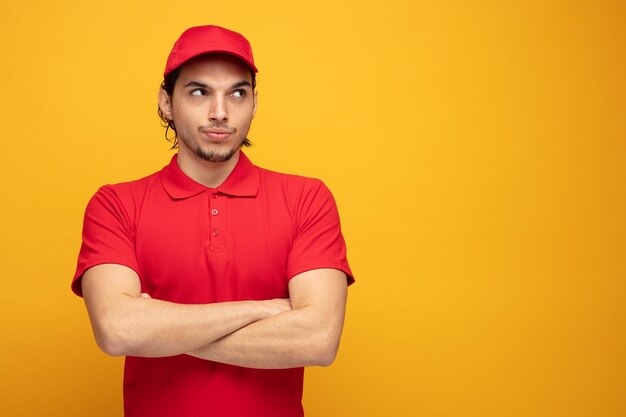 Entregador jovem inseguro vestindo uniforme e boné olhando para o lado, mantendo os braços cruzados isolados em fundo amarelo com espaço de cópia