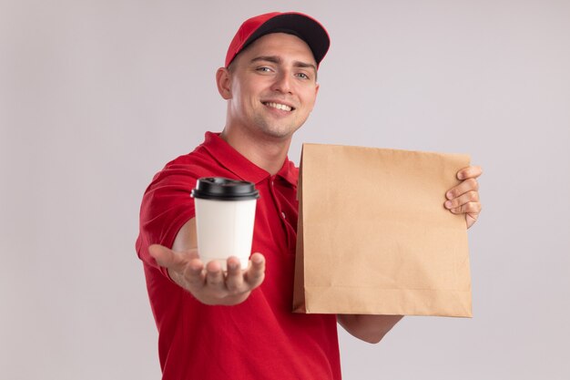 Entregador jovem sorridente, vestindo uniforme com tampa segurando um pacote de comida de papel e segurando uma xícara de café na frente, isolada na parede branca