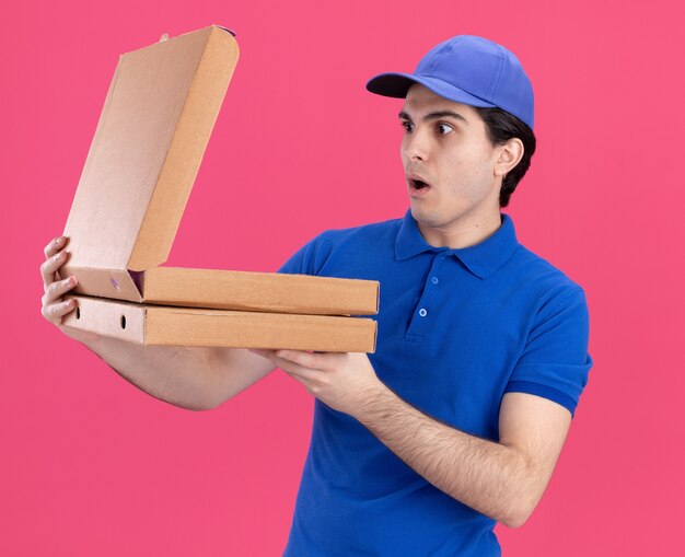Entregador jovem surpreso de uniforme azul e boné segurando pacotes de pizza, abrindo um deles, olhando para dentro, isolado na parede rosa
