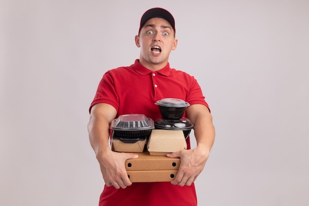 Entregador jovem surpreso vestindo uniforme com tampa segurando recipientes de comida em caixas de pizza isoladas na parede branca