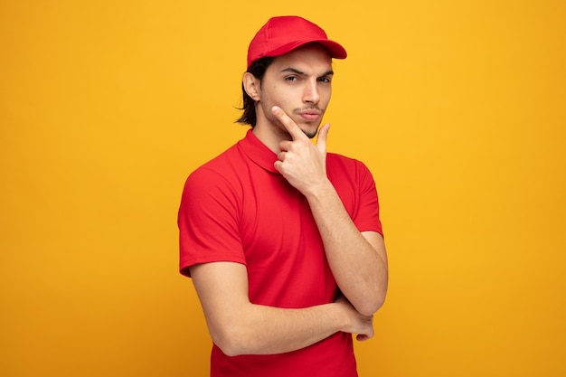 entregador jovem suspeito vestindo uniforme e boné mantendo a mão no queixo olhando para câmera isolada em fundo amarelo