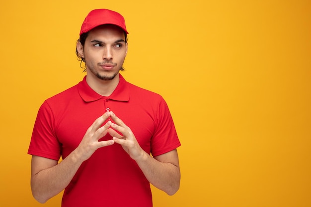 Entregador jovem suspeito vestindo uniforme e boné olhando para o lado enquanto mantém as mãos juntas isoladas em fundo amarelo com espaço de cópia