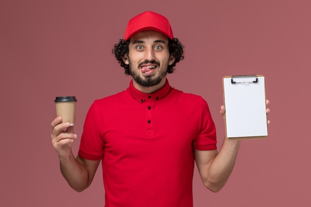 Entregador masculino de camisa vermelha e capa, vista frontal, segurando a xícara de café marrom e o bloco de notas na parede rosa claro funcionário de entrega de uniforme de trabalho