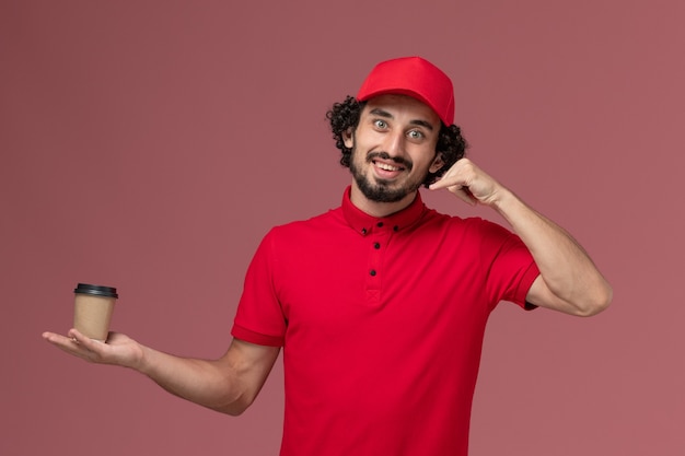 Entregador masculino de camisa vermelha e capa, vista frontal, segurando a xícara de café marrom na parede rosa, entregador uniforme de serviço trabalhador trabalhador