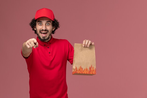 Entregador masculino de camisa vermelha e capa, vista frontal, segurando um pacote de comida na parede rosa funcionário da empresa de entrega de serviço de trabalho masculino