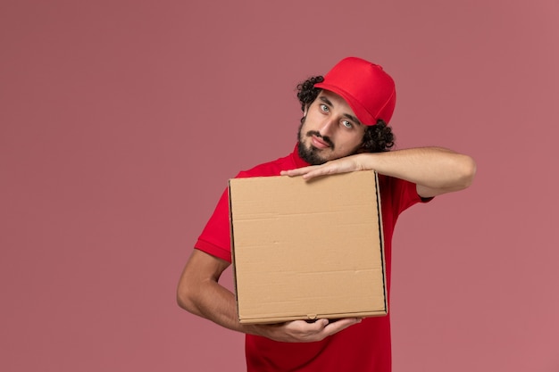 Entregador masculino de camisa vermelha e capa, vista frontal, segurando uma caixa de entrega de comida sobre o funcionário da empresa de entrega de serviço de mesa rosa