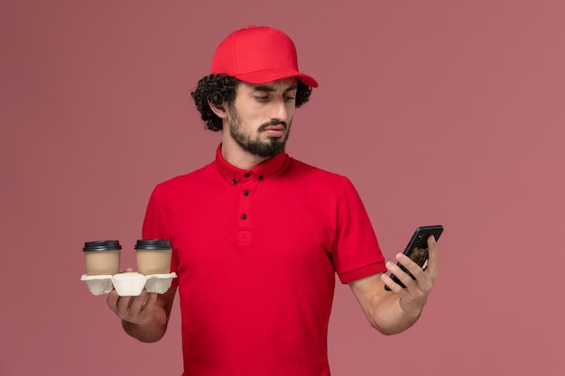 Entregador masculino de camisa vermelha e capa, vista frontal, segurando xícaras de café marrons e telefone na parede rosa claro funcionário de entrega de serviço masculino