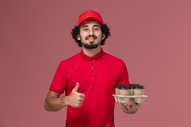 Entregador masculino de camisa vermelha e capa, vista frontal, segurando xícaras de café marrons na parede rosa claro funcionário de trabalho de entrega de serviço