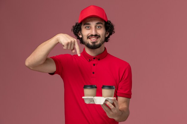 Entregador masculino de camisa vermelha e capa, vista frontal, segurando xícaras de café marrons na parede rosa serviço de entrega de funcionário trabalhador.