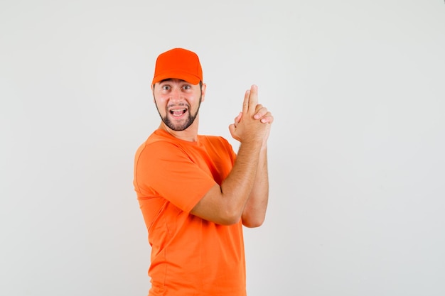 Entregador mostrando gesto de arma em t-shirt laranja, boné e olhando alegre, vista frontal.