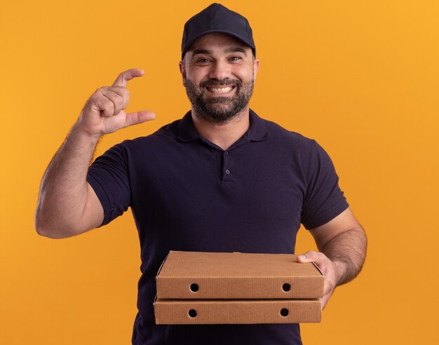 Entregador sorridente de meia-idade de uniforme e boné segurando caixas de pizza mostrando o tamanho isolado na parede amarela