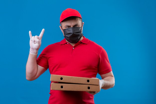 Entregador, vestindo uniforme vermelho e boné na máscara protetora facial segurando caixas de pizza, fazendo o símbolo do rock com cara séria sobre parede azul