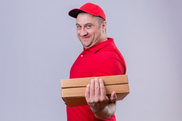 Entregador vestindo uniforme vermelho e boné segurando caixas de pizza sorrindo amigável com cara feliz sobre parede branca isolada