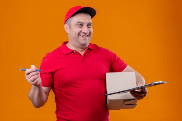 Entregador vestindo uniforme vermelho e boné segurando o pacote caixa e prancheta com caneta olhando de lado com cara feliz sorrindo por cima da parede laranja