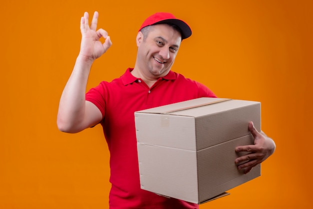 Entregador vestindo uniforme vermelho e boné segurando o pacote caixa olhando positivo e feliz fazendo ok assinar parede laranja